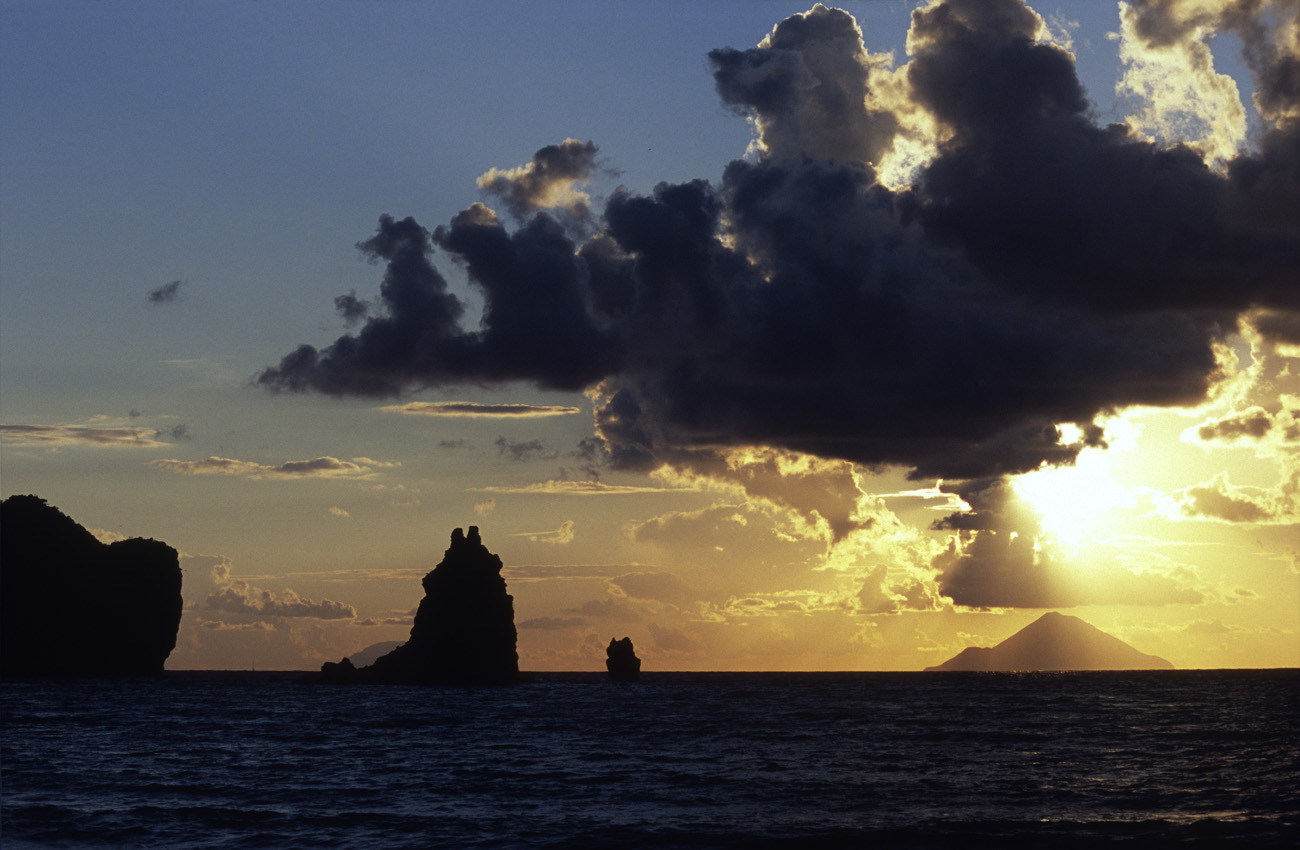 eol06_129.jpg - Italie, îles Eoliennes Vulcano coucher de soleil depuis la Spiaggia Sabbia Nera: au fond Filicudi, qu'un caprice du hasard a coiffée d'un nuage donnant à penser qu'il s'agit des fumées d'une éruption Fuji Velvia 100 F