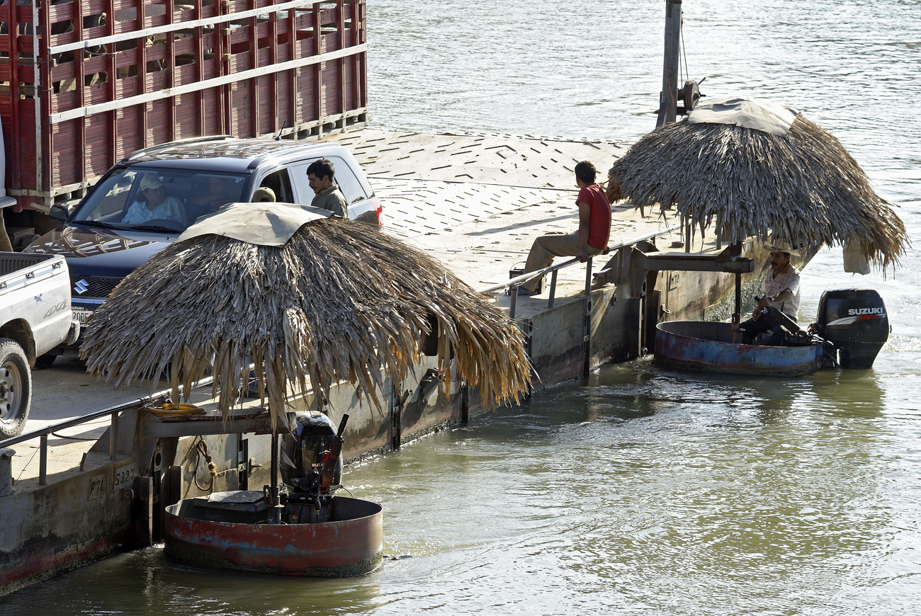 gua06_1131.jpg - rio de la Passion  Sayaxché le bac du rio de la Passion, transportant de gros véhicules avec peu de moyens : deux moteurs hors bord et deux toits de paille pour protéger ceux qui les maneuvrent..