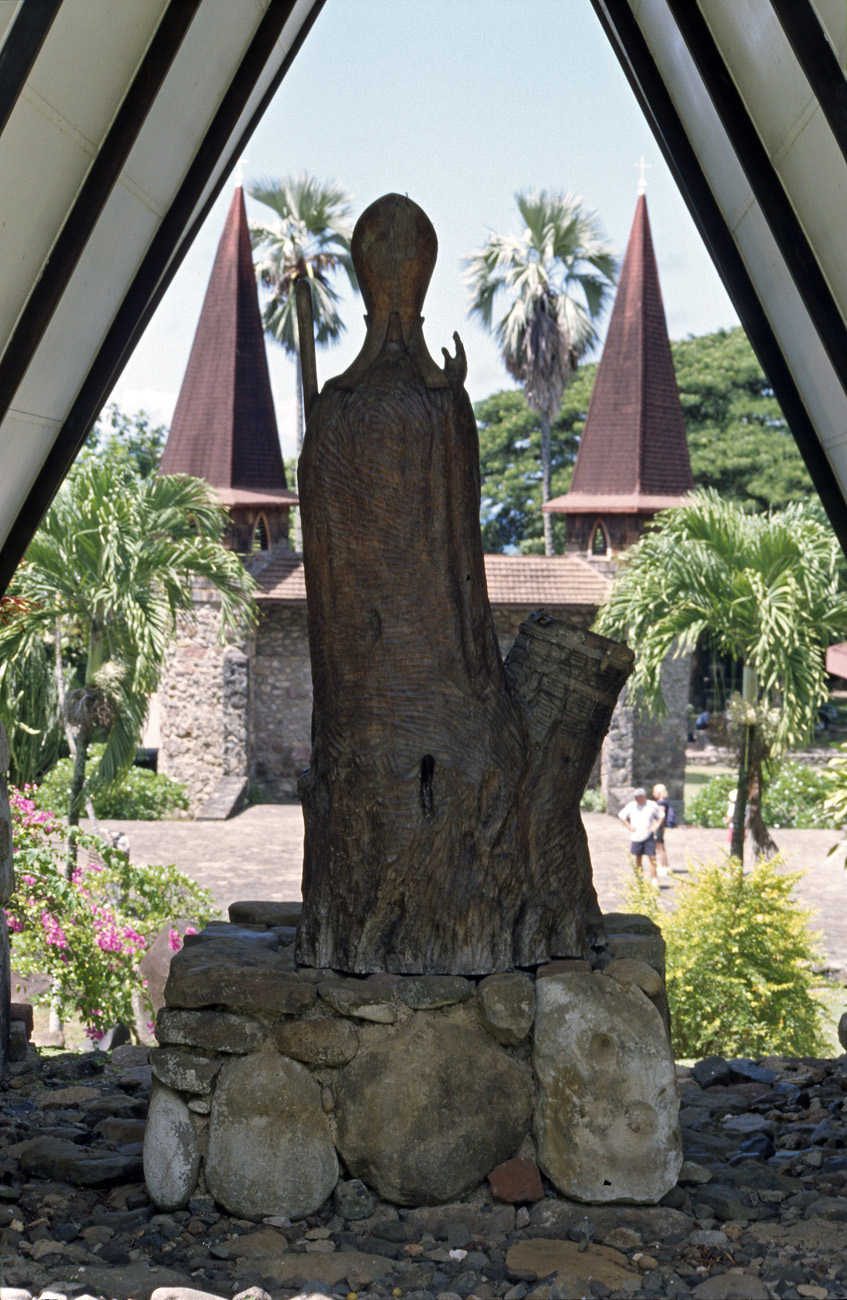 pol05_0265m.jpg - Iles Marquises, Nuku Hiva Taiohae : en fond, les tours de l'ancienne église, au premier plan la statue de monseigneur Dordillon, sculptée dans un tronc d'arbre